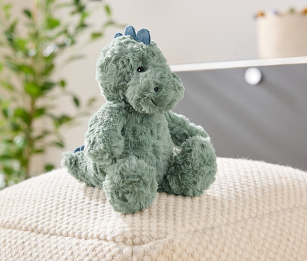 Zelený plyšový dinosaurus sedí na bílé strukturované stoličce. V pozadí je rostlina a skříň.