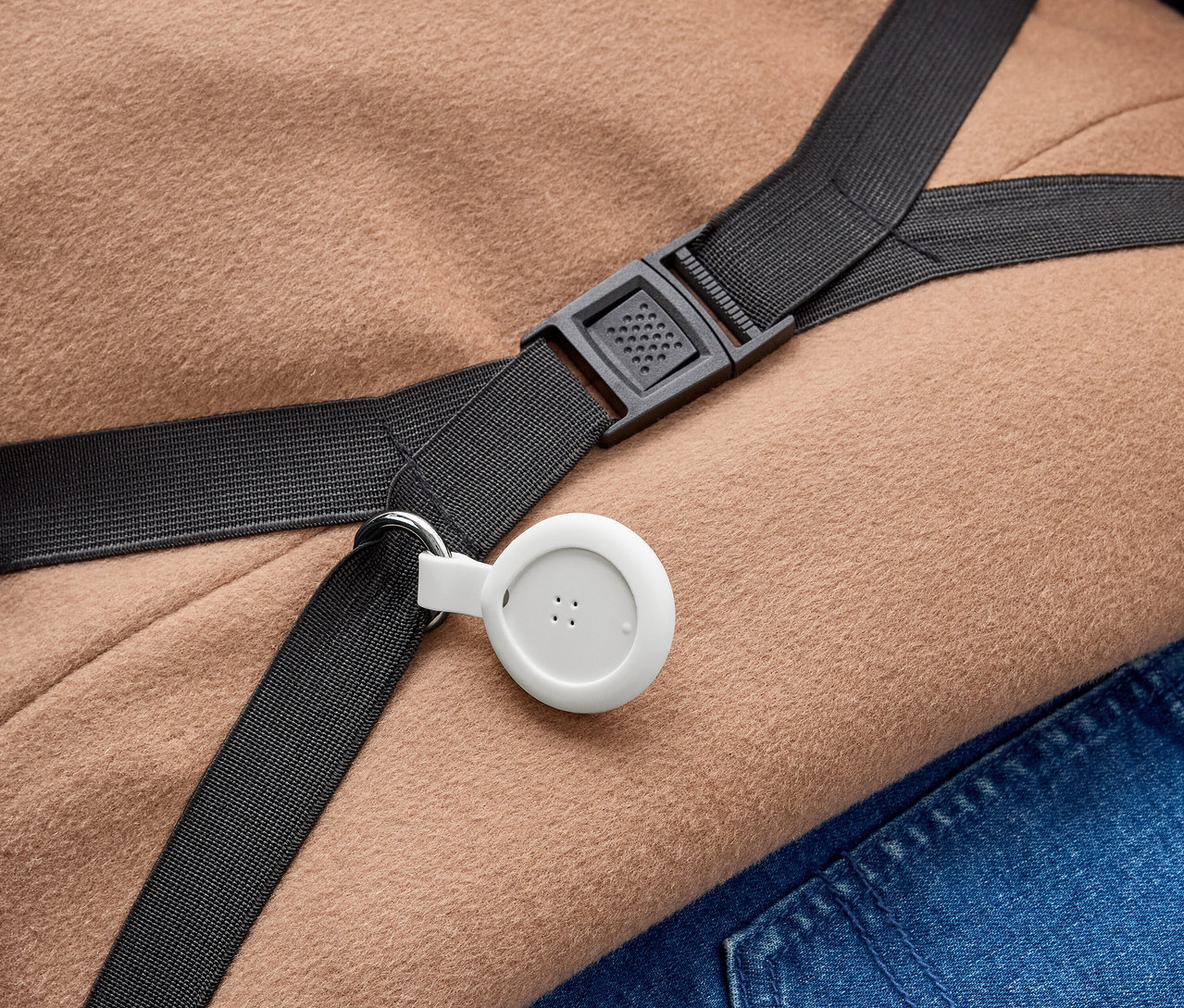 Bílé, kruhové zařízení se třemi tečkami je připevněno k černému popruhu s kovovým kroužkem. Popruh s černou plastovou sponou leží na světle hnědé látce a modré džínovině.