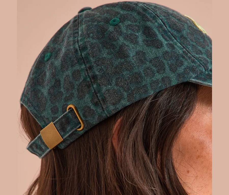 Detail osoby s tmavě zelenou kšiltovkou CECIL s leopardím vzorem a zlatou sponou. Jsou vidět hnědé vlasy.