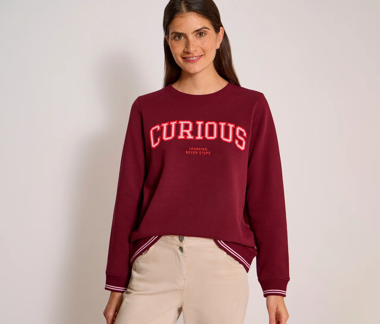 Usměvavá žena s tmavými vlasy má na sobě bordovou mikinu CECIL s nápisem „CURIOUS“ v červenobílých písmenech a „LEARNING NEVER STOPS“ pod ním, v kombinaci se světle béžovými kalhotami.