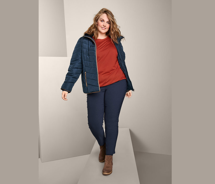 Žena kráčí po molu a má na sobě prošívanou bundu, triko s krátkým rukávem a výšivkou a bengalínové kalhoty.