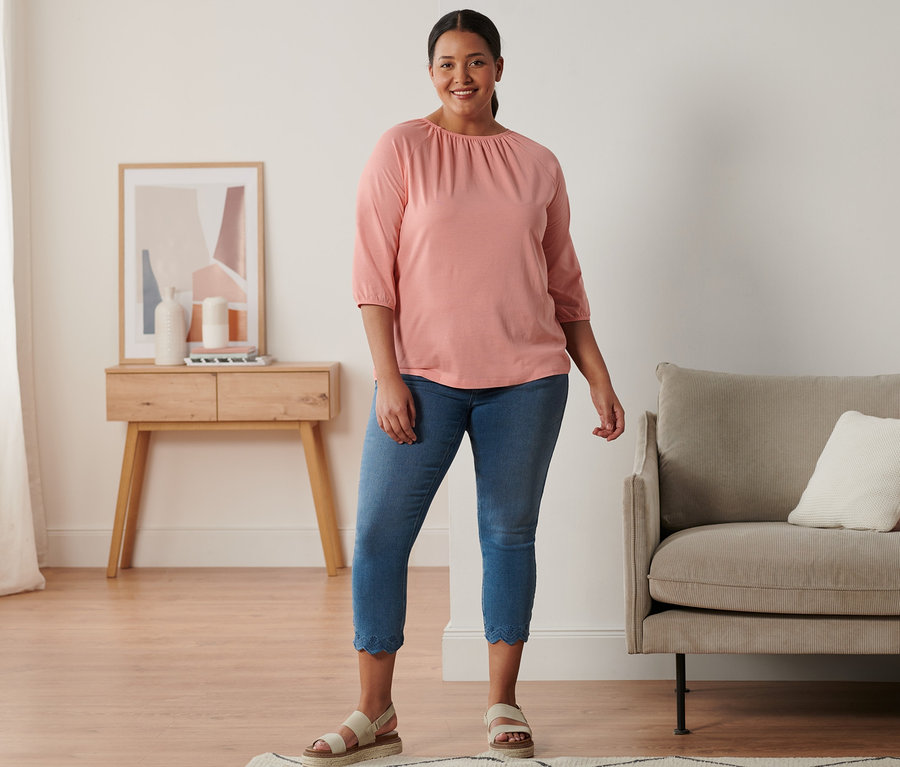 Žena stojí a má na sobě kožené sandály na platformě, 7/8 tregíny s ažurovou výšivkou a korálové triko s 3/4 rukávem.