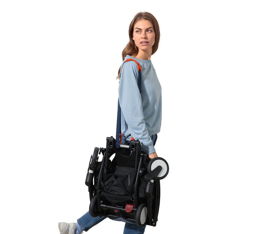 Žena nese složený černý kočárek přes rameno pomocí multifunkčního popruhu.