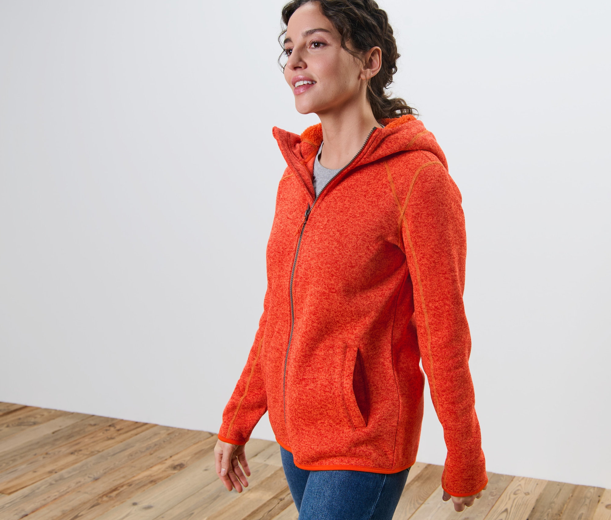 Žena má na sobě oranžovou melírovanou bundu z pleteného fleecu s kapucí.