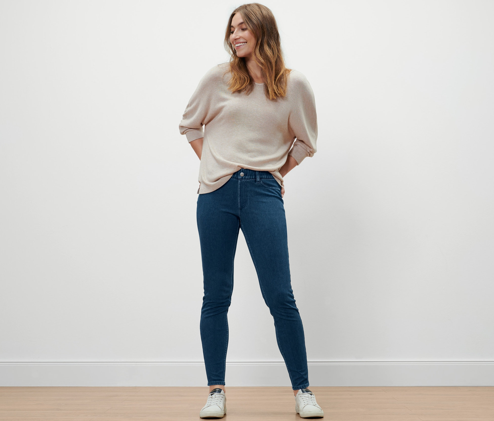 Žena pózuje v džegínách, svetru a teniskách.