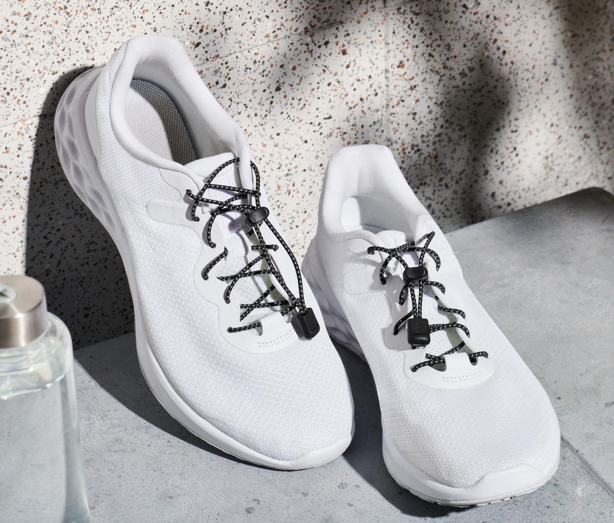 Dvoje bílé tenisky s elastickými tkaničkami a láhev s vodou.