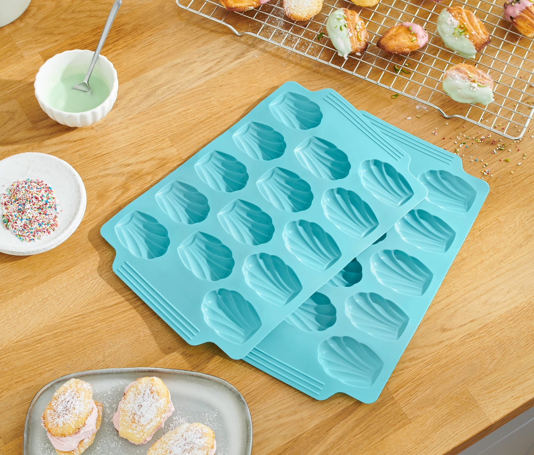 Na dřevěném stole leží 2 ks forem na pečení madlenek, upečené madlenky, cukrářské posypky a polevy.