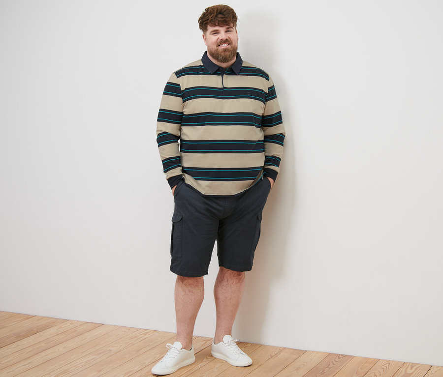 Muž stojí a má na sobě polo tričko s dlouhým rukávem, kraťasy a tenisky.