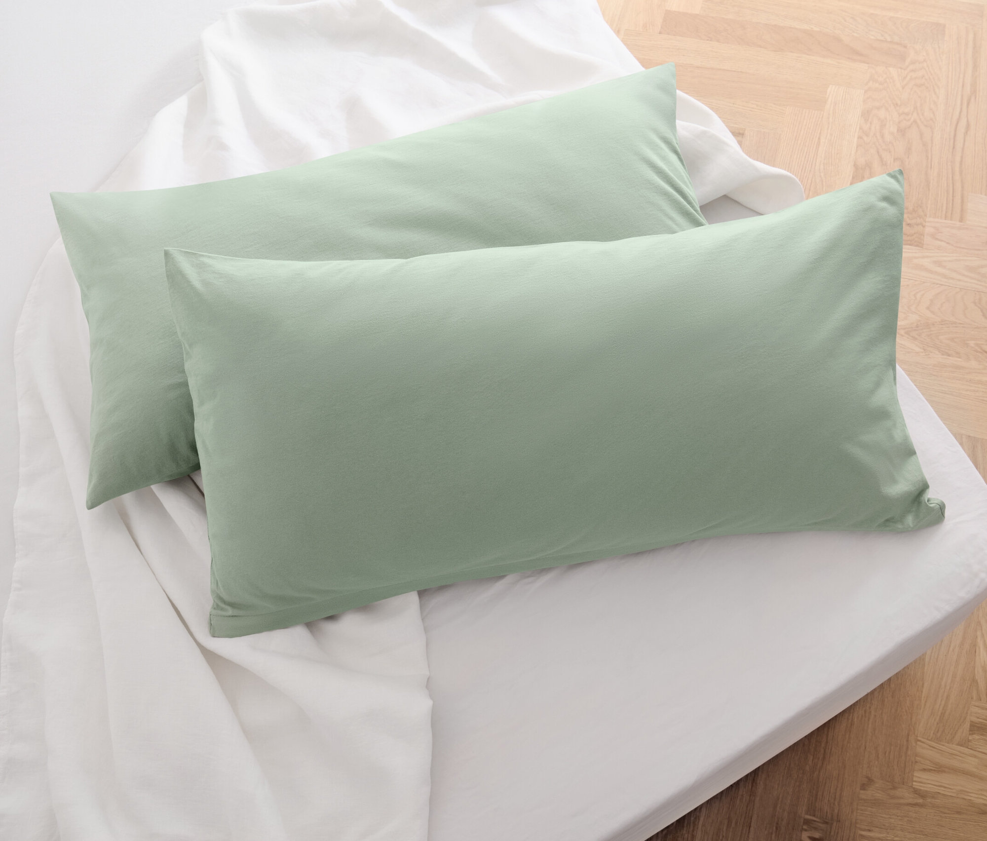 Dva žerzejové povlaky na polštáře šalvějově zelené barvy leží na bílém prostěradle.