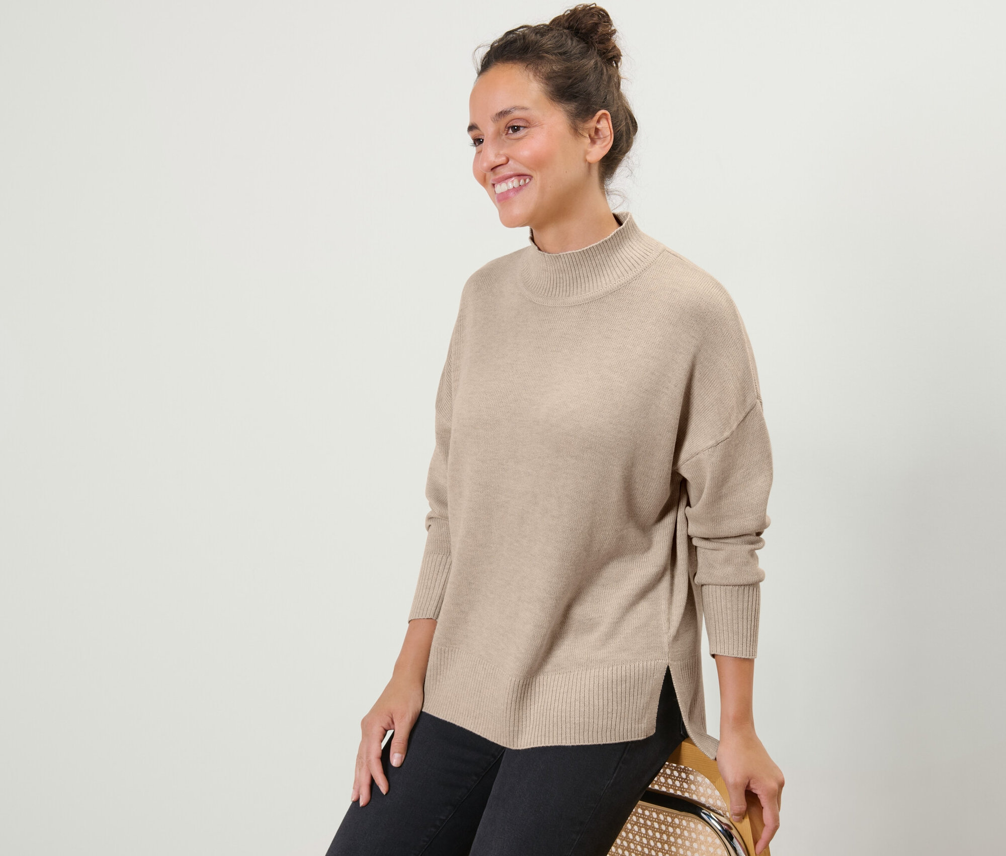 Usmívající se žena sedí a má na sobě béžový svetr z jemného úpletu s rolákem a černé džíny.