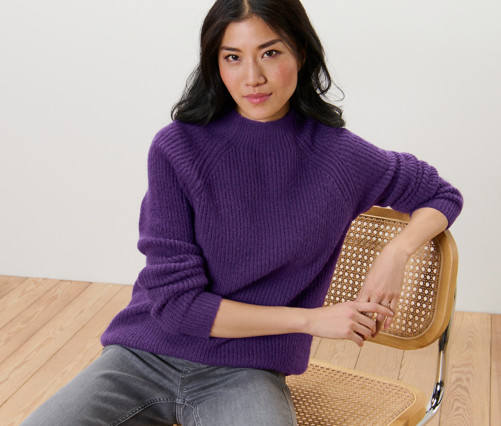 Žena sedí na židli a má na sobě fialový svetr z hrubého úpletu se stojáčkem.