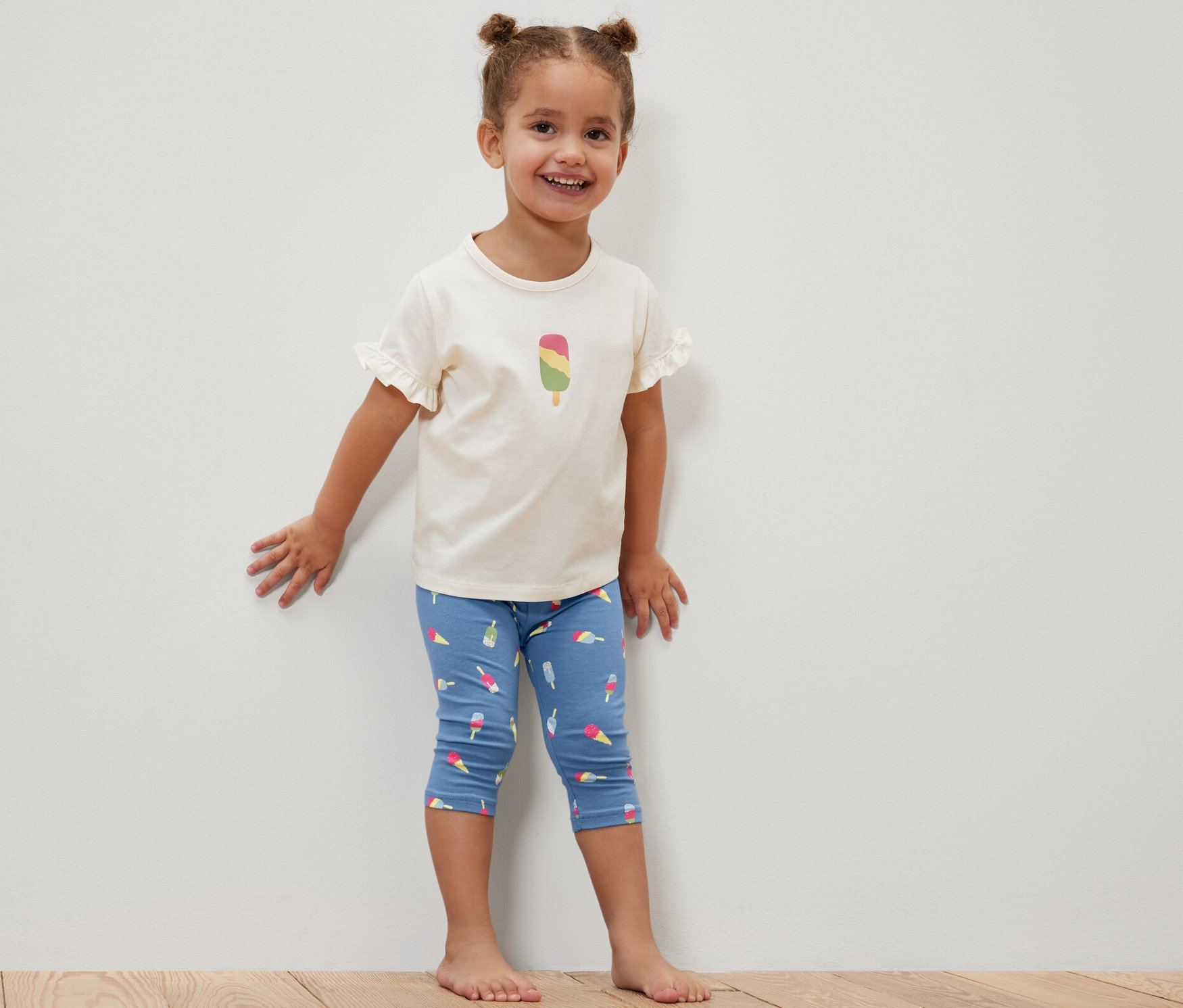 Usmívající se holčička stojí u bílé stěny a má na sobě modré tříčtvrteční legíny s potiskem nanuků a bílé tričko s obrázkem nanuku. Dětské ¾ legíny, 2 ks, modrá.