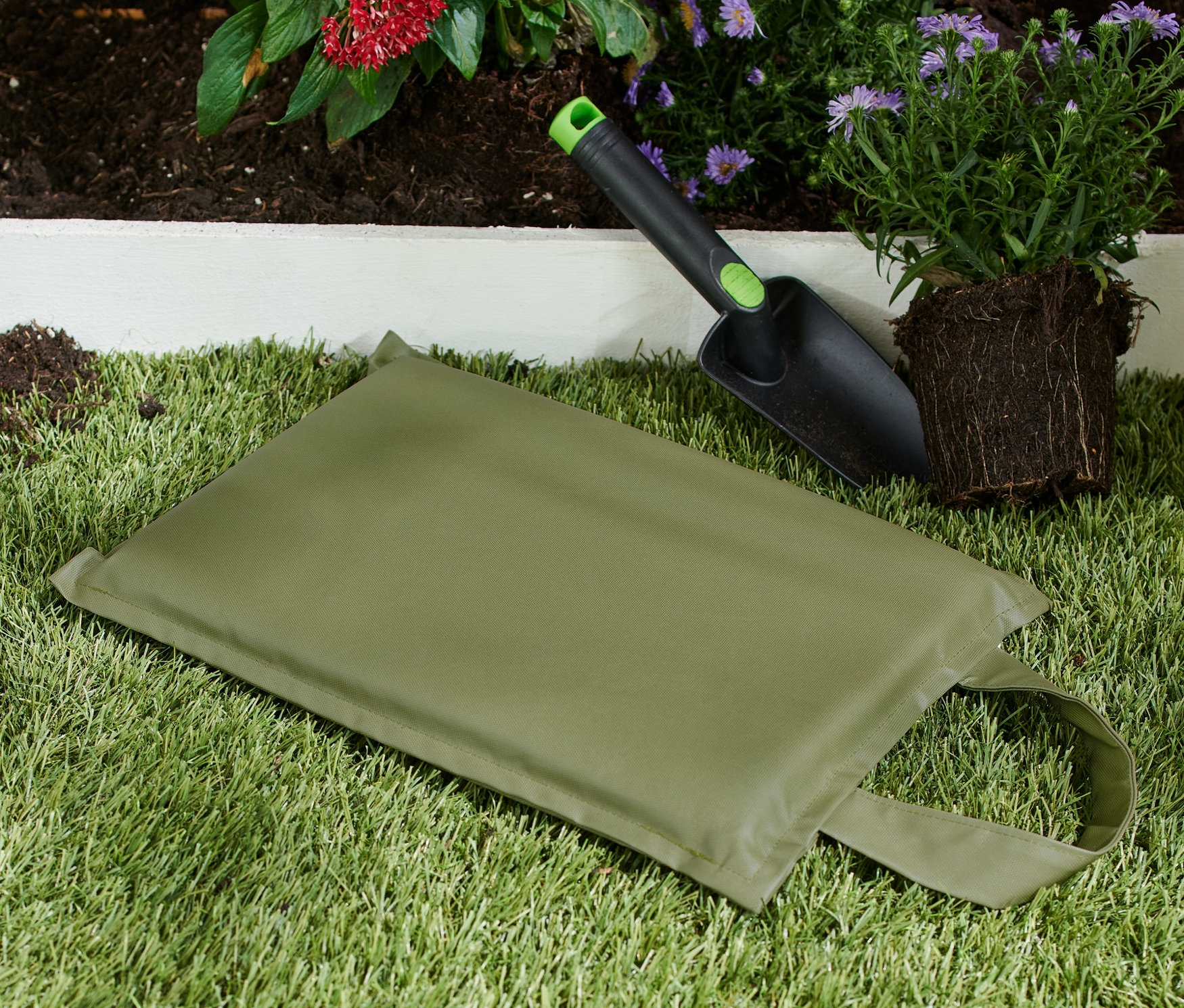 Zahradnický polštářek na klečení leží na trávě vedle lopatky a rostliny.