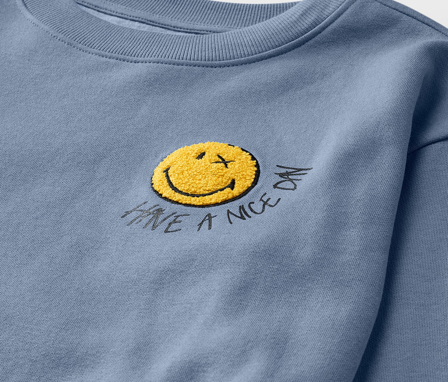 Detail světle modré dětské mikiny SMILEYWORLD® s vnitřní stranou s broušeným povrchem a žlutým smajlíkem.