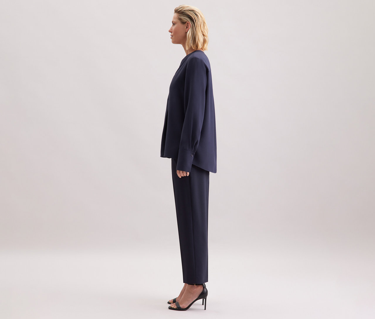 Žena stojí bokem, má na sobě tmavě modrou tuniku z viskózy Seidensticker, kalhoty a černé sandály.