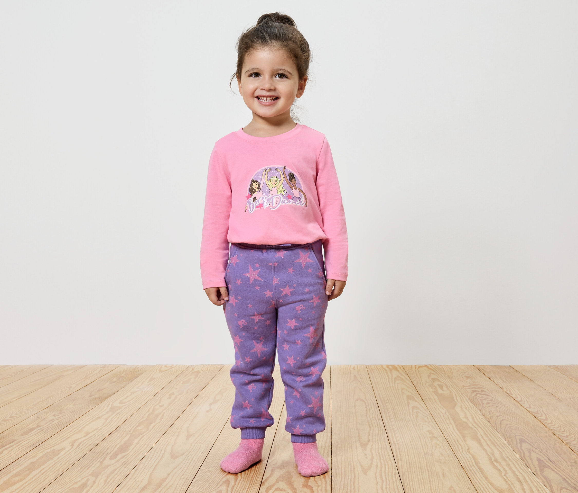 Usmívající se holčička pózuje v růžovém tričku s potiskem a fialových kalhotách s hvězdami.