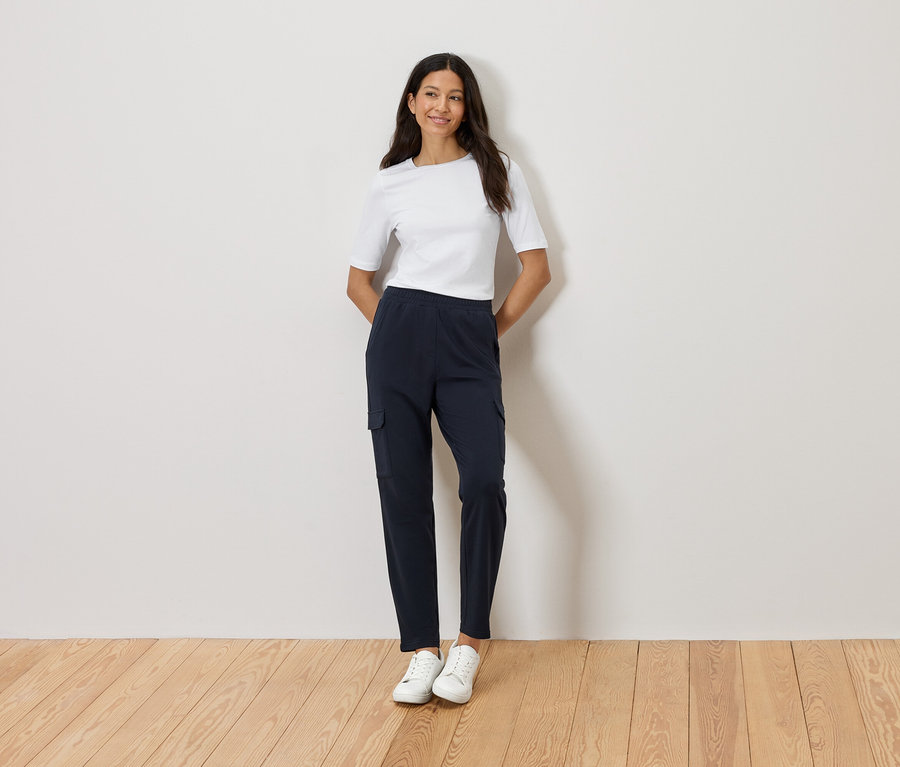 Žena stojí se založenýma rukama před zdí, má na sobě bílé tričko, tmavě modré cargo kalhoty a bílé tenisky.