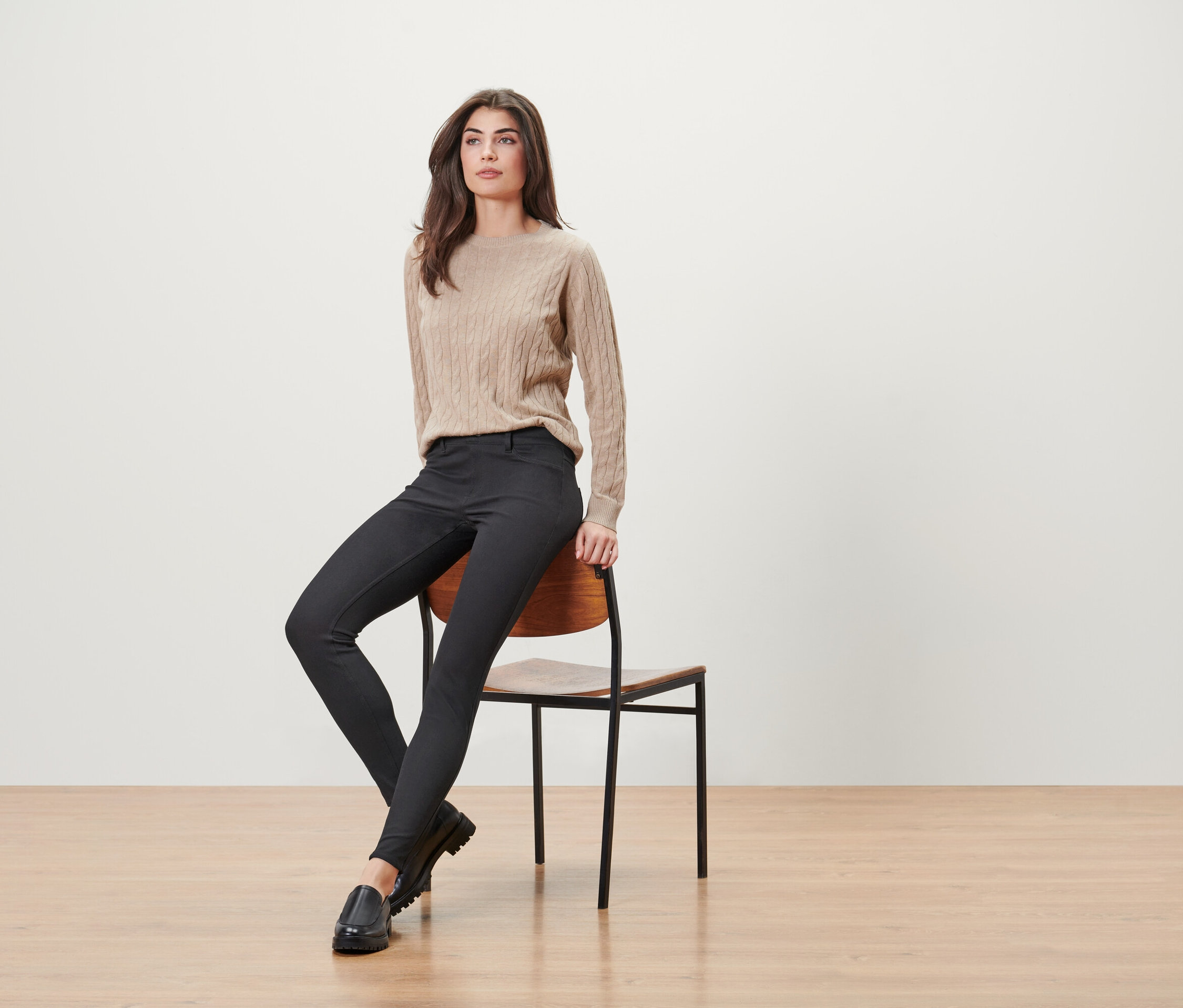 Žena sedí na dřevěné židli, má na sobě béžový svetr, černé kalhoty a černé mokasíny.