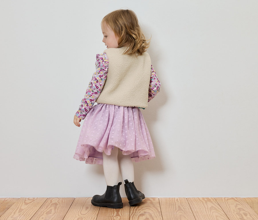 Malá holčička stojí zády ke kameře, má na sobě halenku s květinovým vzorem, světlou vestu, fialovou sukni a černé boty.