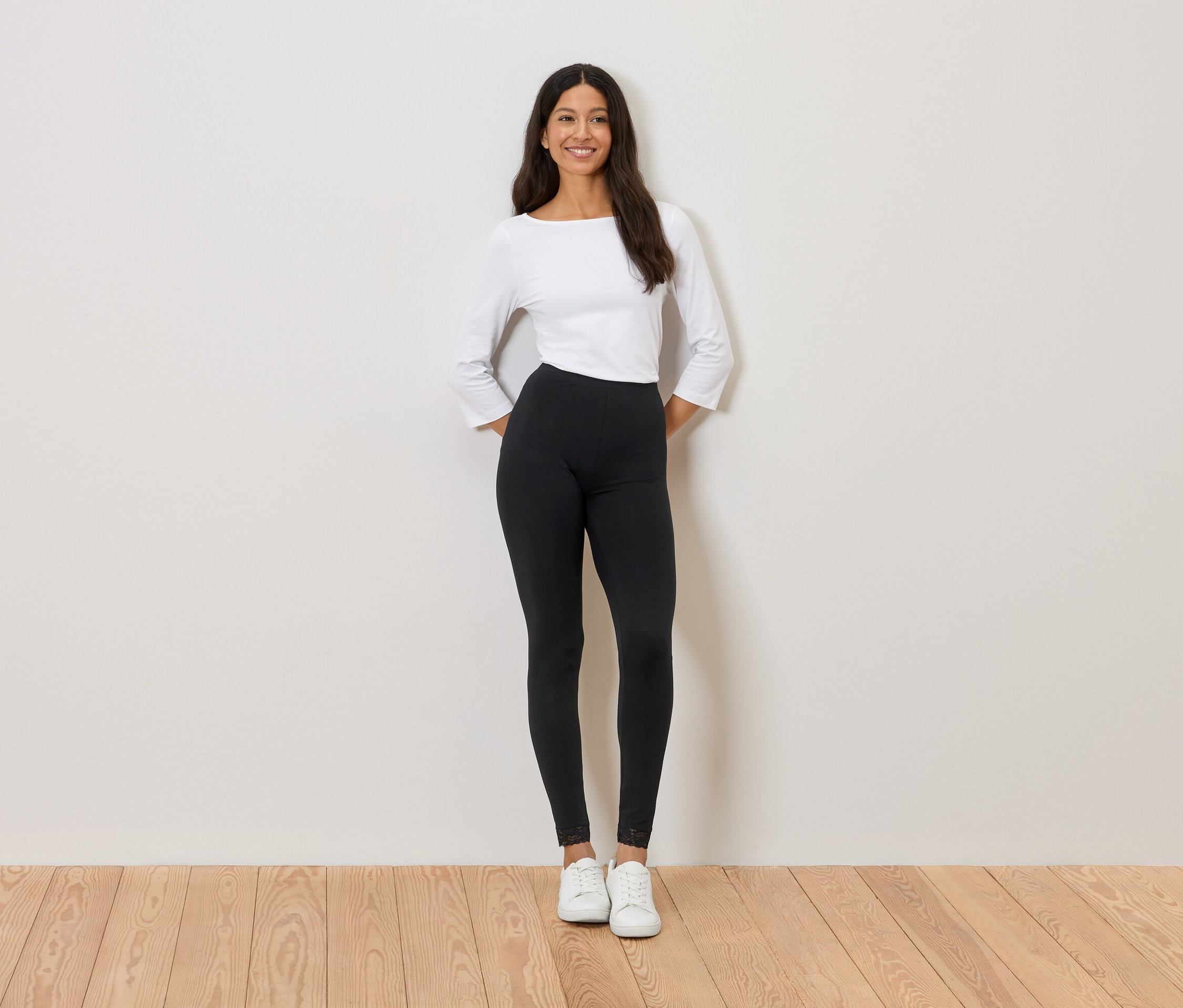 Žena stojí před stěnou, má na sobě bílý top, černé legíny a bílé tenisky.