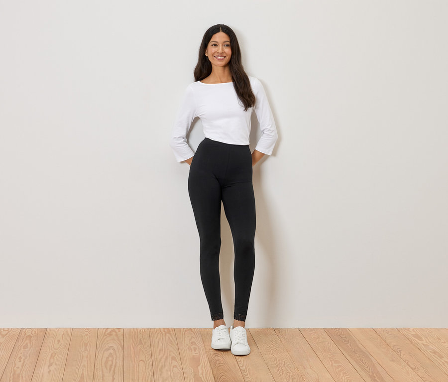 Žena stojí před stěnou, má na sobě bílý top, černé legíny a bílé tenisky.