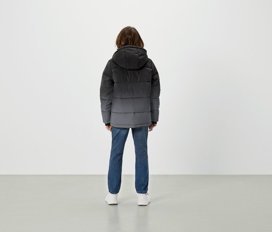 Zezadu je vidět osoba v šedé péřové bundě s kapucí, modrých džínách a bílých botách.