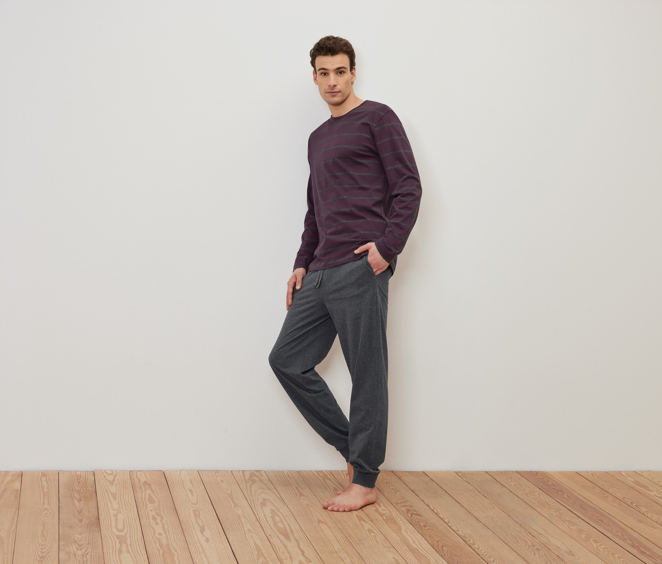 Muž stojí bosý na dřevěné podlaze a má na sobě fialové pruhované tričko s dlouhým rukávem a šedé tepláky.