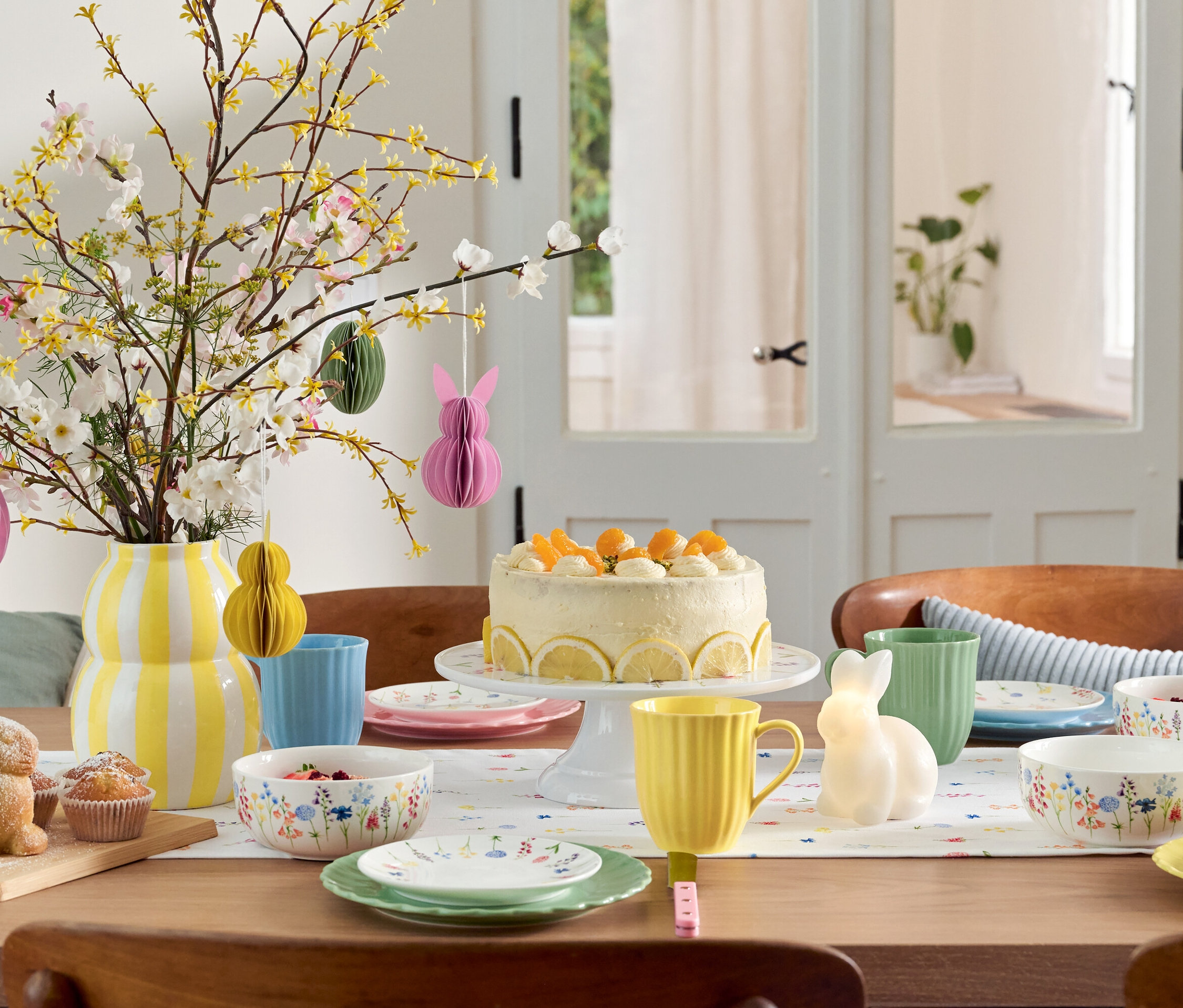 Jídelní stůl s velikonoční výzdobou, dortem, nádobím a vázou s květinami.