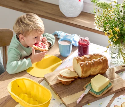 Blonďatý chlapec jí chleba s džemem u stolu s vánočkou, máslem, džemem, nádobím a květinami.