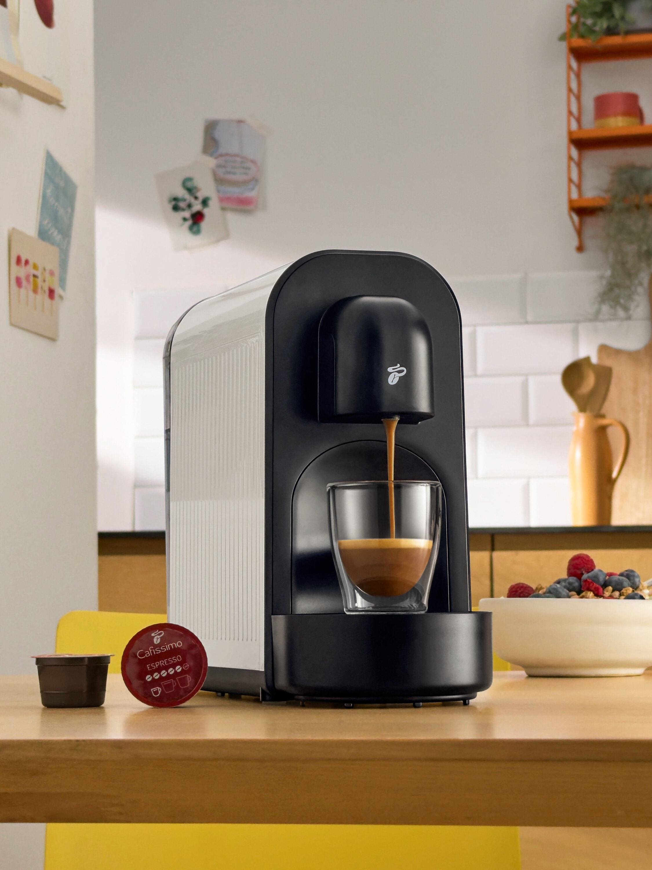 Černý kávovar nalévá kávu do sklenice. Vedle něj leží kávové kapsle a miska s bobulemi.