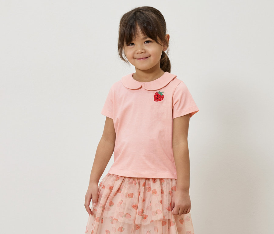 Holčička v růžovém tričku s aplikací jahody a růžové sukni s jahodovým vzorem.