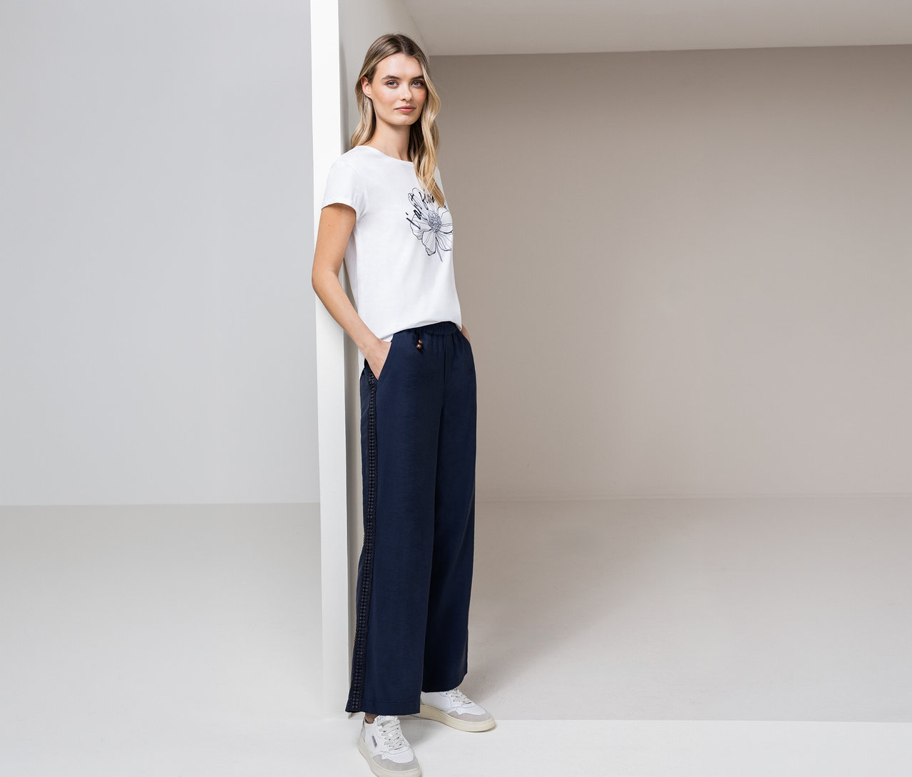 Žena pózuje ve světlé místnosti v tričku Street One, modrých kalhotách a bílých teniskách.