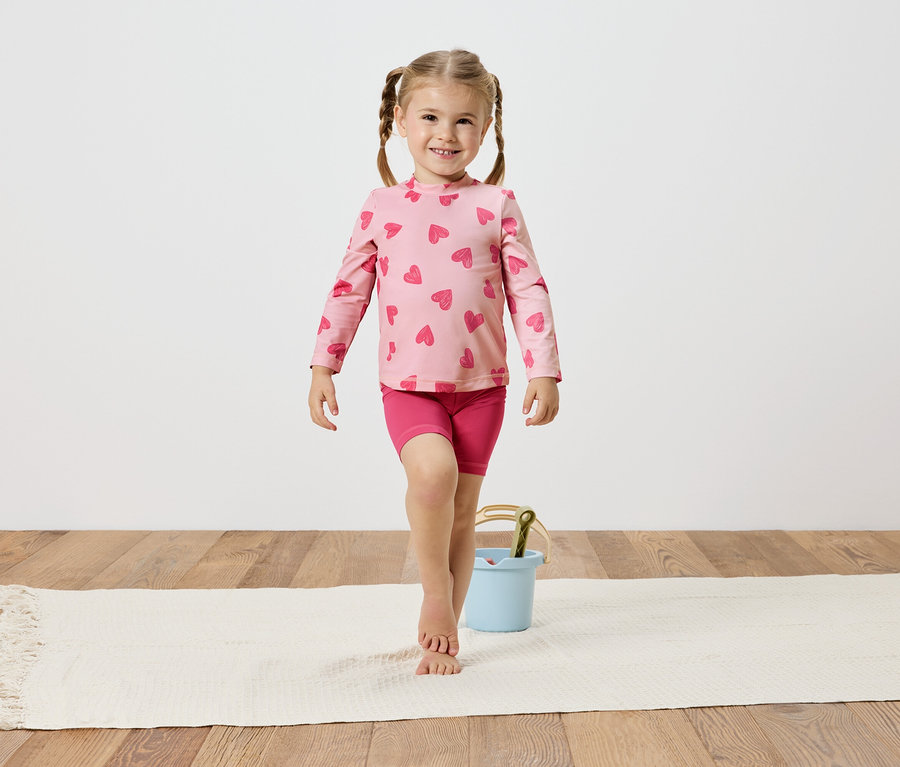 Dívka s copánky v růžovém topu se srdíčky a šortkách balancuje na bílém koberci vedle světle modrého kbelíku.