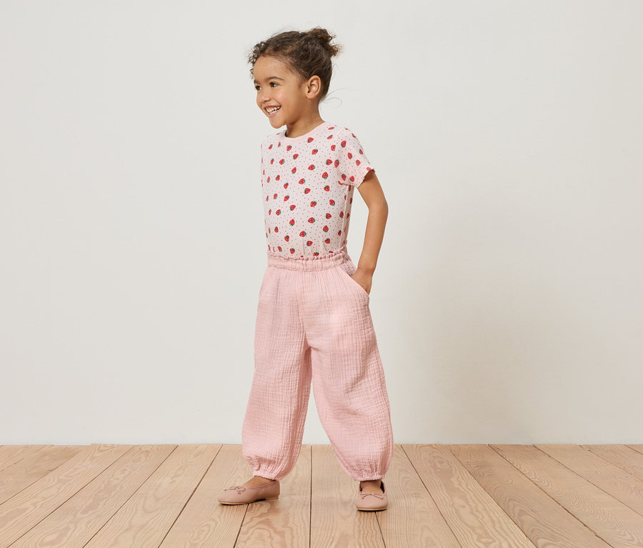 Dívka stojí na dřevěné podlaze v růžovém jahodovém tričku a růžových kalhotách.