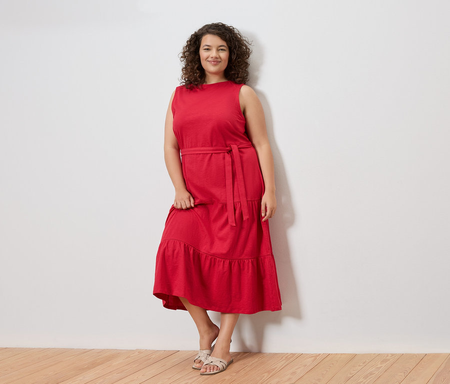 Žena s kudrnatými vlasy stojí před bílou zdí a má na sobě červené šaty bez rukávů s páskem a sandály.