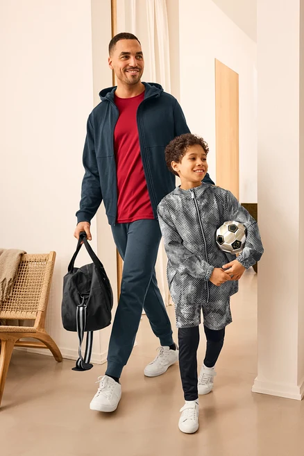 Muž se sportovní taškou a dítě s fotbalem, oba mají na sobě sportovní oblečení. Muž má na sobě Sweatjacke a Sweathose. Dítě má na sobě Kinder-Windbreaker-Jacke.
