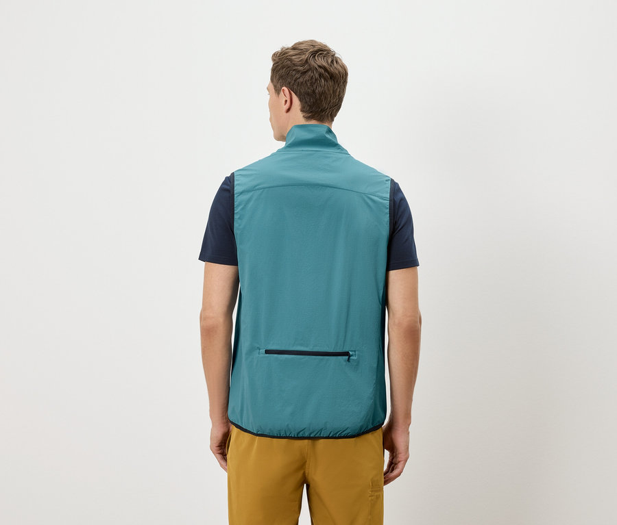 Muž zezadu v námořnickém tričku a zlatých šortkách, má na sobě petrolejovou vestu s kapsou na zip.