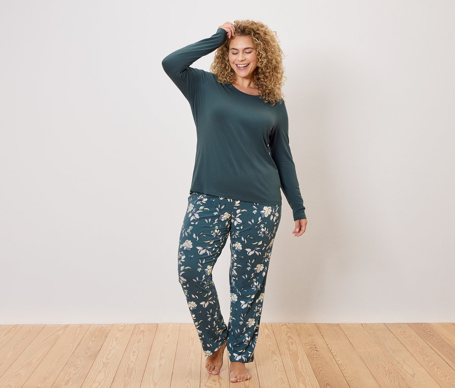 Žena s kudrnatými vlasy má na sobě tmavě zelený top s dlouhým rukávem a tmavě zelené pyžamové kalhoty s květinovým vzorem.