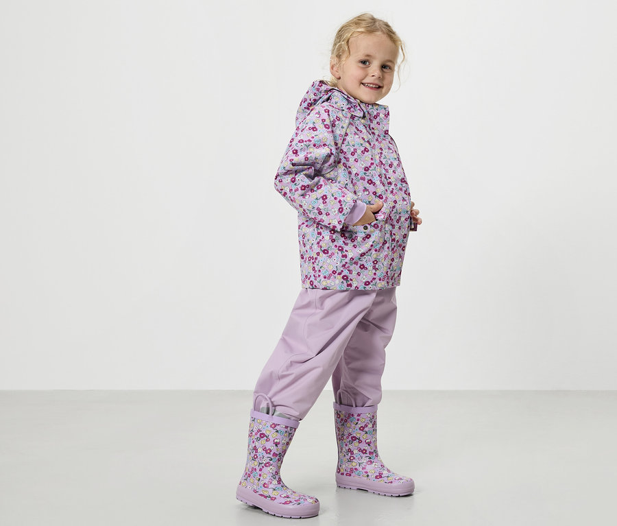 Blonďatá dívka má na sobě fialový plášť do deště s květinovým vzorem, fialové kalhoty do deště a odpovídající gumáky.
