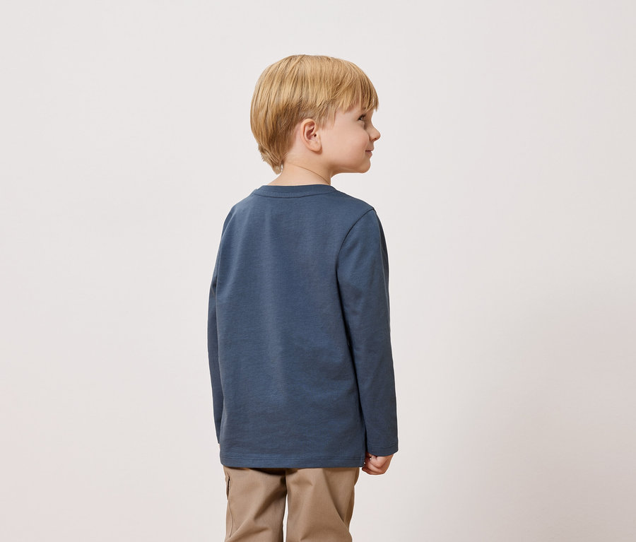 Blonďatý chlapec stojí zády k fotoaparátu a má na sobě modré tričko s dlouhým rukávem a khaki kalhoty.