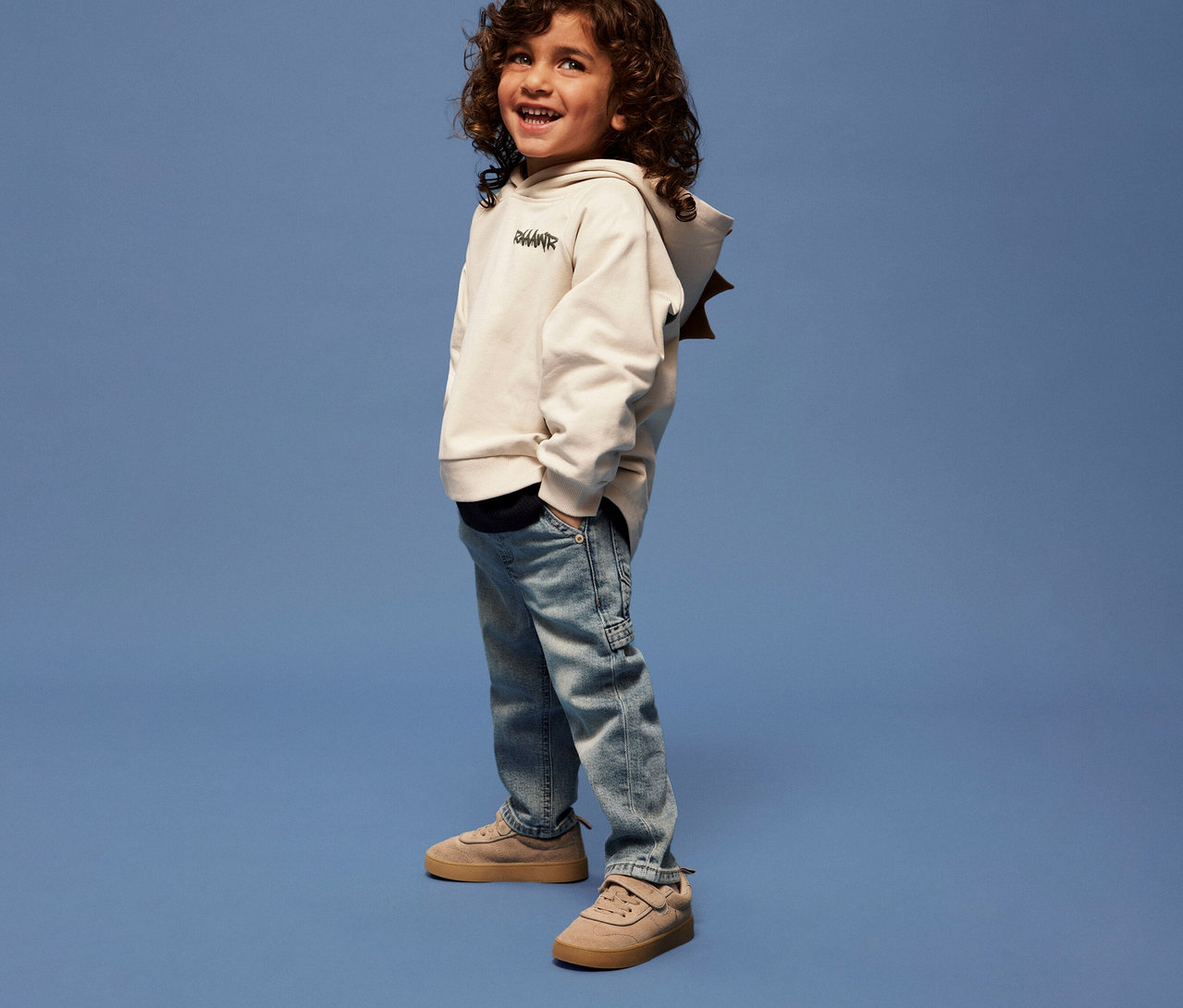 Chlapec s kudrnatými vlasy má na sobě dětskou mikinu s 3D dinosauří aplikací NAME IT, džíny a tenisky na modrém pozadí.