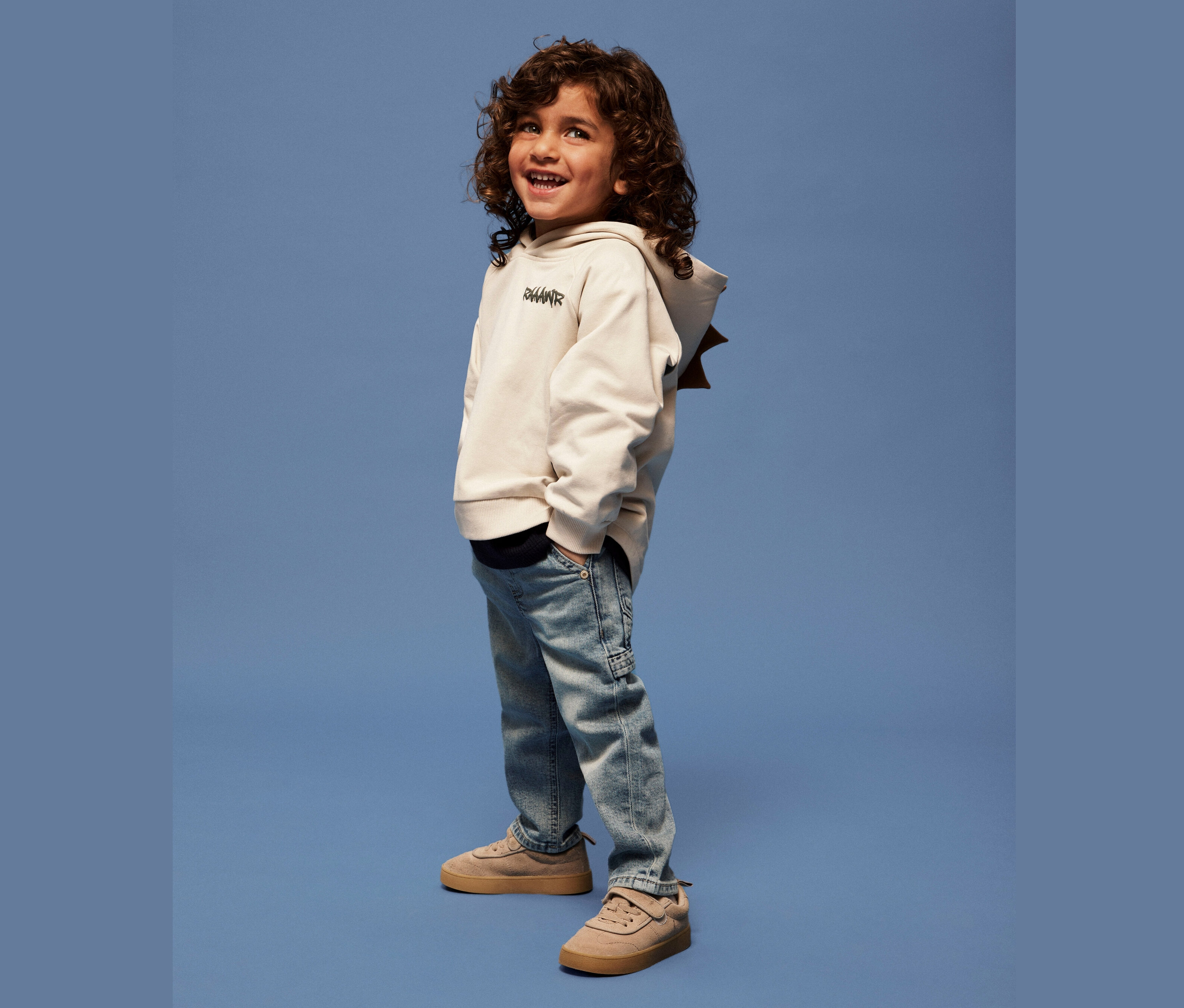 Usmívající se chlapec s kudrnatými vlasy stojí na modrém pozadí v béžové mikině s kapucí, světle modrých džínách a hnědých botách.