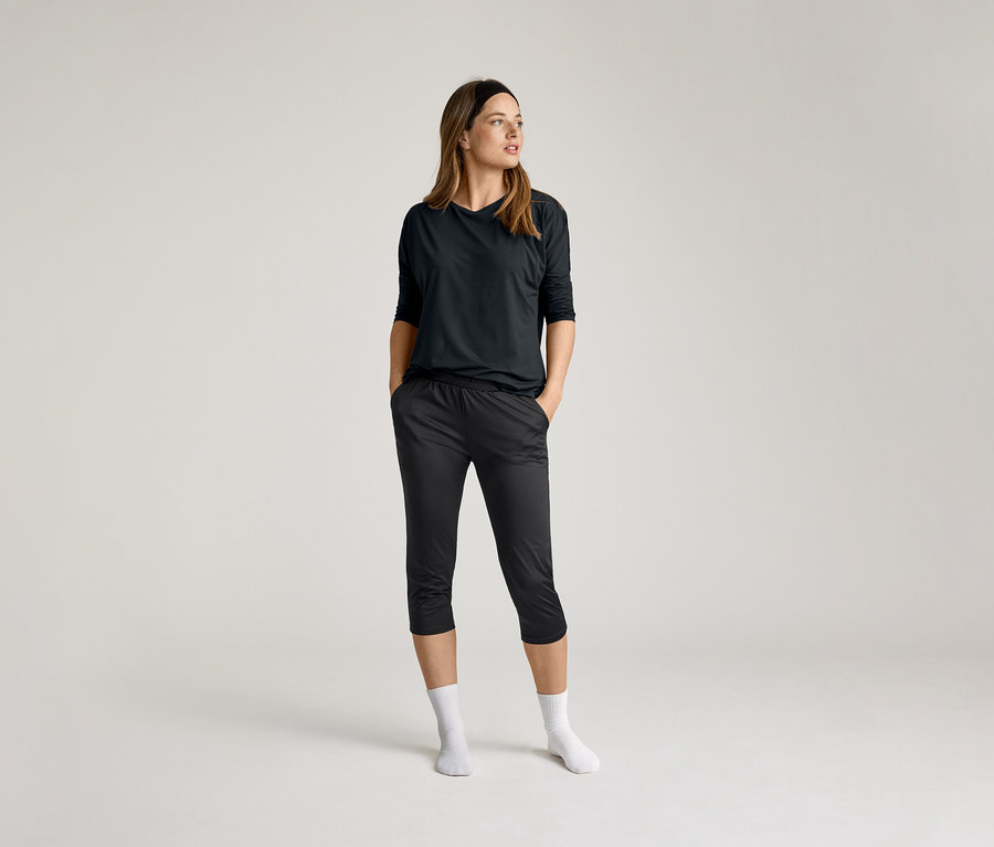 Žena v černém 3/4 sportovním tričku a tmavých kalhotách, ruce v kapsách, bílé ponožky, na světlém pozadí.
