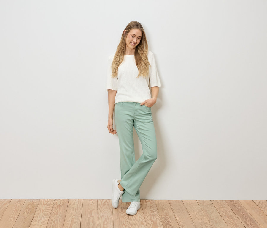 Žena s blond vlasy v bílém topu, zelených kalhotách a bílých teniskách stojí na dřevěné podlaze před bílou stěnou.