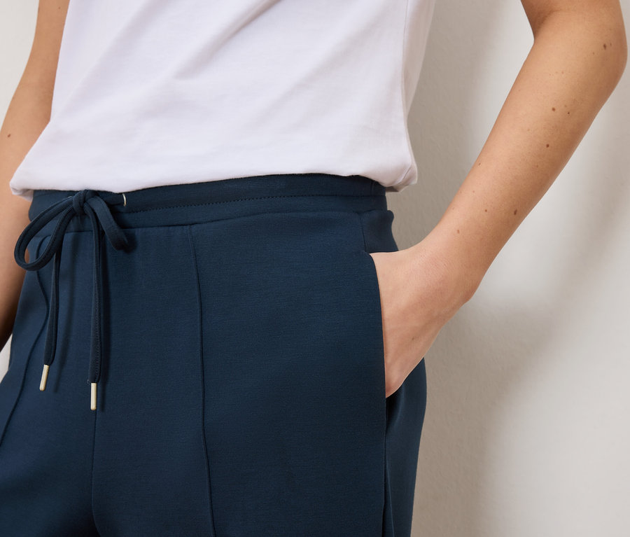 Detailní záběr na ženu v tmavě modrých teplácích se stahovací šňůrkou a zlatými detaily a bílém tričku.