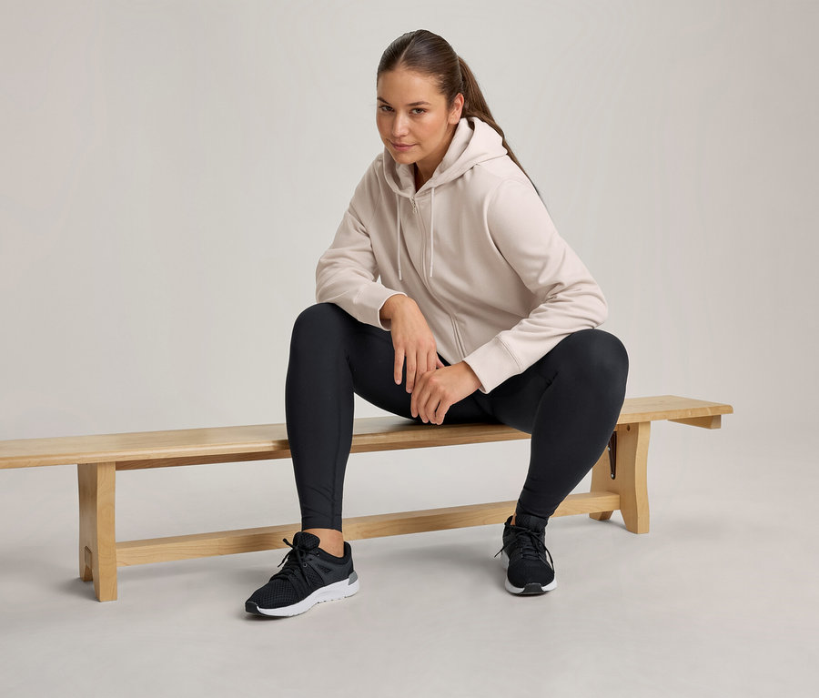 Žena sedí na dřevěné lavičce, má na sobě světle béžovou mikinu na zip s kapucí, černé legíny a černé sportovní boty.