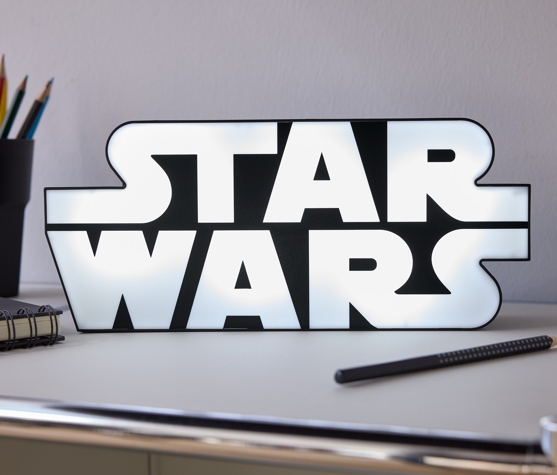 Na stole stojí svítící nápis Star Wars, vedle něj je tužka, zápisník a kelímek s pastelkami.