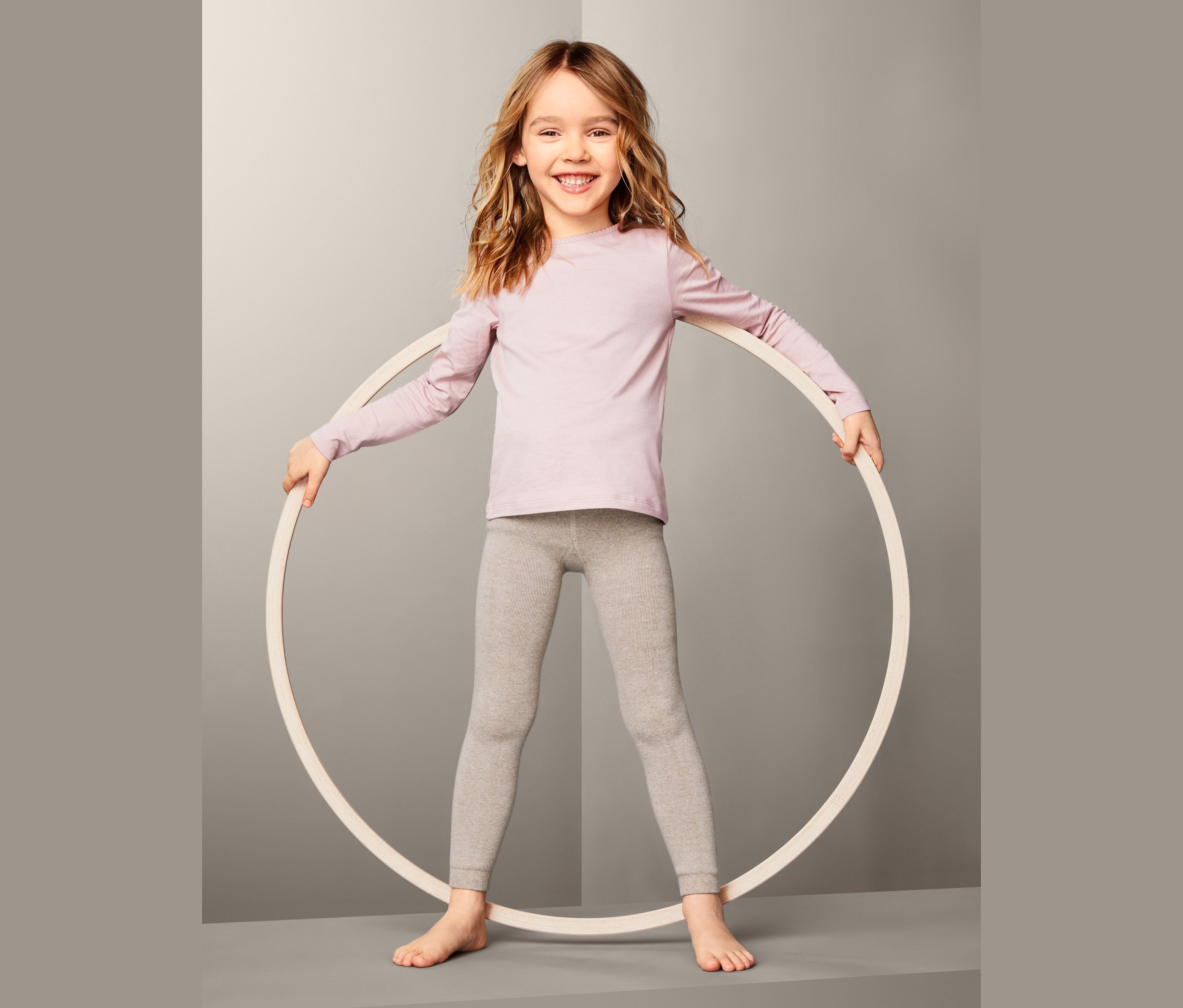 Usmívající se dívka drží kruh. Má na sobě růžové tričko a šedé termo legíny, 2 ks.