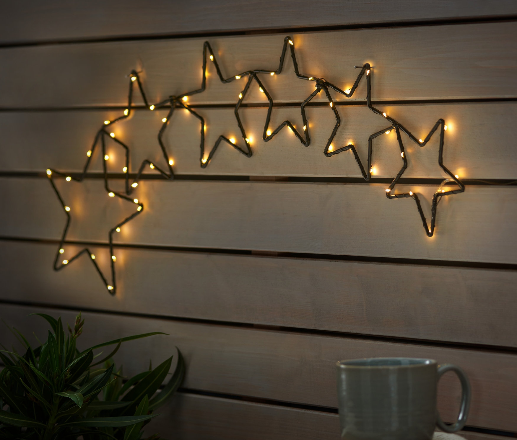 Na dřevěné stěně visí LED hvězdy. Na dřevěném stole stojí hrnek a talíř s hvězdami.