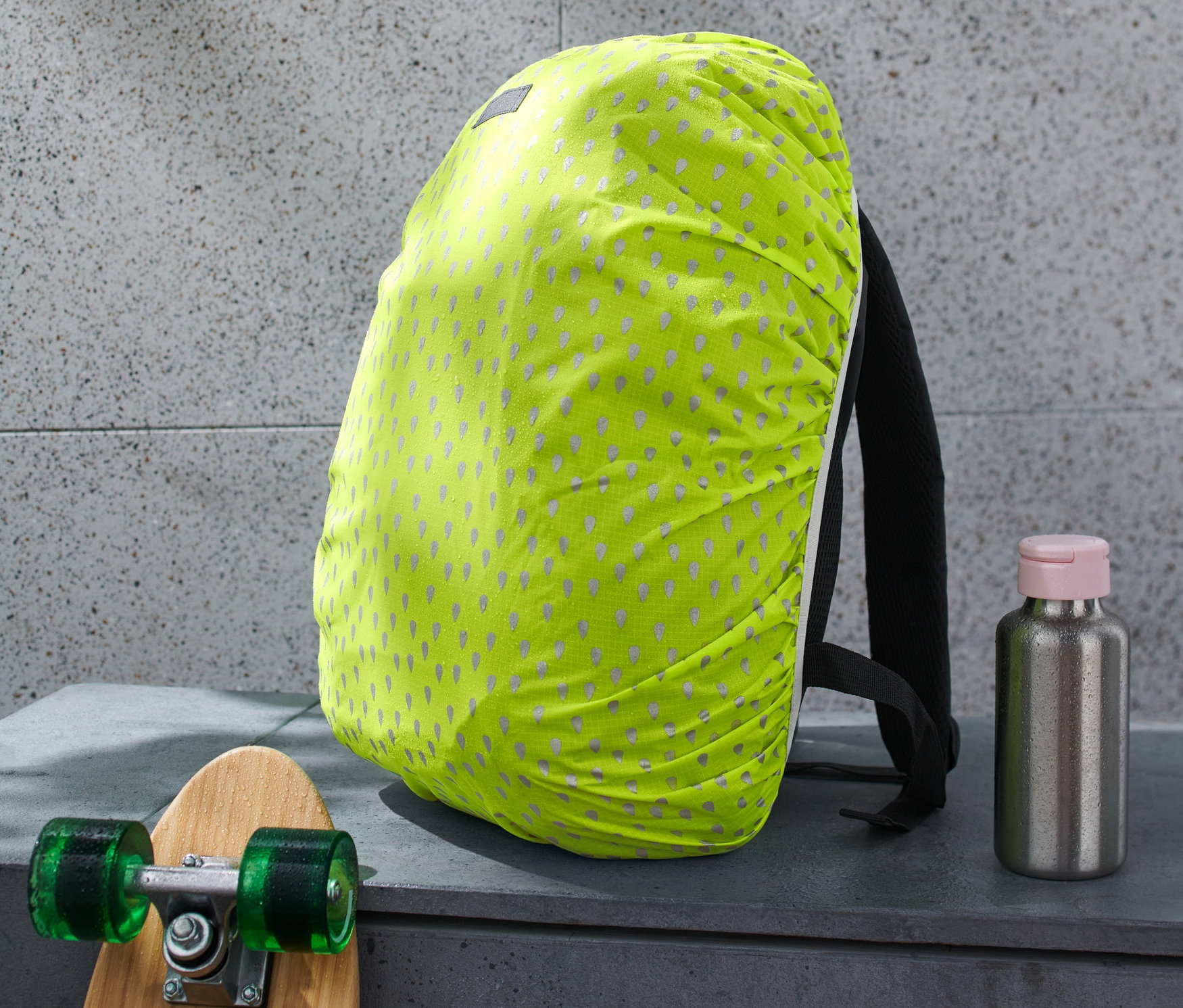 Batoh s reflexní pláštěnkou, vedle skateboard a láhev na pití.